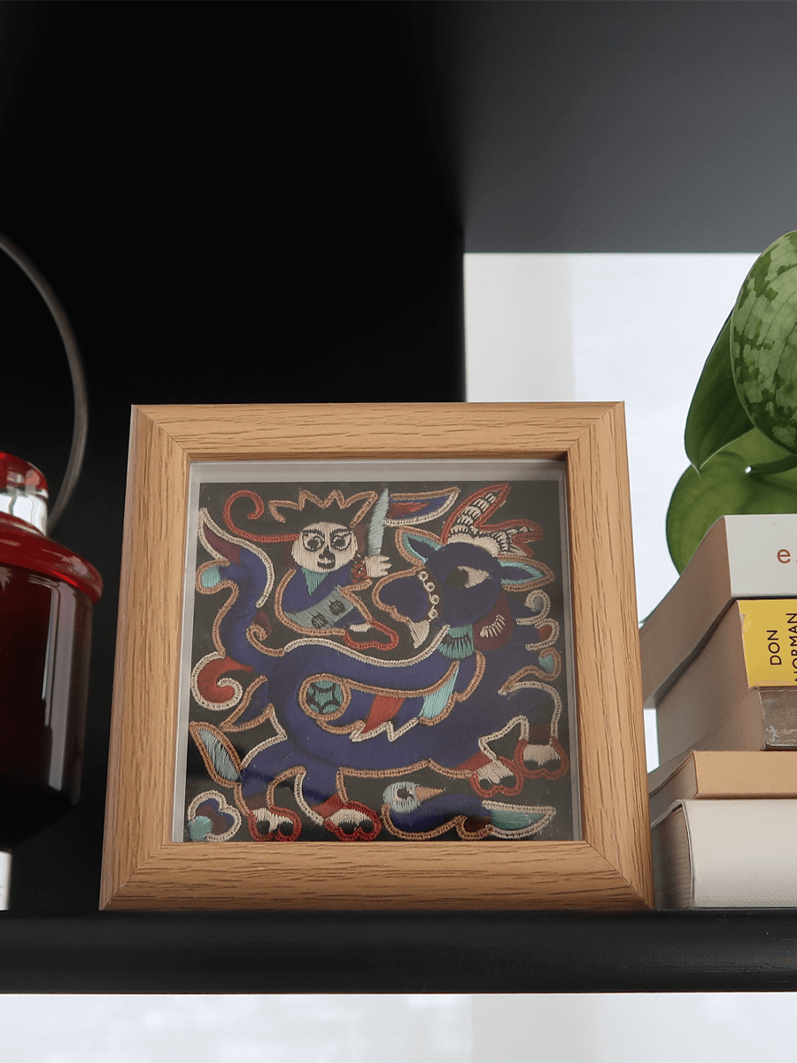 Framed textile art with a cultural and traditional design on a shelf with books and a plant.